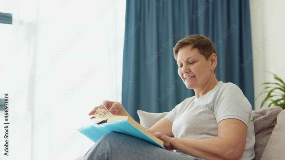 Relaxed senior woman sitting on sofa and reading book indoors at home. Leisure activity of mature female at comfort modern flat. Casual elderly life and wrinkled skin closeup Relaxed senior woman sitting on sofa and reading book indoors at home. Leisure activity of mature female at comfort modern flat. Casual elderly life and wrinkled skin closeup