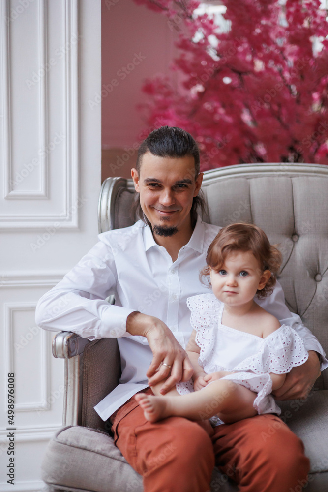 Fototapeta premium Handsome happy father sitting with his little daughter on beige sofa indoors. Young man in white shirt with his 1-year-old daughter in white dress, holding her in hands on legs. family tenderness