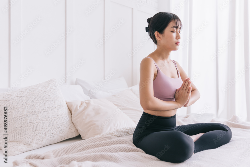 Young serene attractive sporty Asian woman practicing yoga on the bed, doing Ardha Padmasana exercise, meditating in Half Lotus pose with namaste, indoor working out at home, wearing sportswear. Young serene attractive sporty Asian woman practicing yoga on the bed, doing Ardha Padmasana exercise, meditating in Half Lotus pose with namaste, indoor working out at home, wearing sportswear.