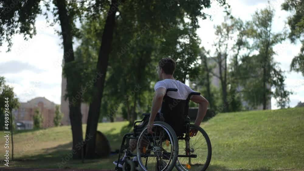 Tracking shot side view of young man pushing wheelchair wheels riding fast in sunbeam. Determined motivated Caucasian sportsman training exercising outdoors in park. Disability and sport concept Tracking shot side view of young man pushing wheelchair wheels riding fast in sunbeam. Determined motivated Caucasian sportsman training exercising outdoors in park. Disability and sport concept