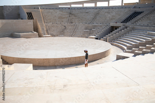 Man in a smart casual, with a hat and looking far, is standing outdoors in an amphitheater