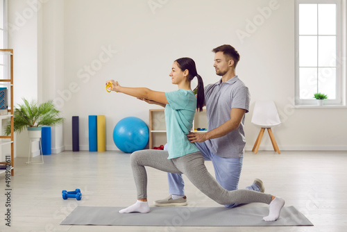 Male physiotherapist helping his female patient do special physical exercises. Side view of happy young woman doing forward lunges on rubber mat at physiotherapy clinic or rehabilitation center