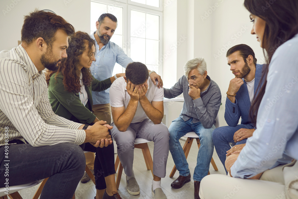 People comforting a grieving crying man. Concerned understanding patients in group therapy supporting a distraught desperate devastated guy who's hiding face in hands and shedding tears of self pity People comforting a grieving crying man. Concerned understanding patients in group therapy supporting a distraught desperate devastated guy who's hiding face in hands and shedding tears of self pity