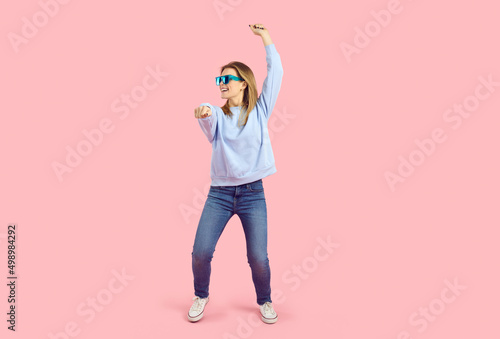 Lively young girl in good mood having fun in studio. Full body shot of happy pretty Caucasian woman in comfortable wear and cool sunglasses doing funny dancing move on pastel pink colour background