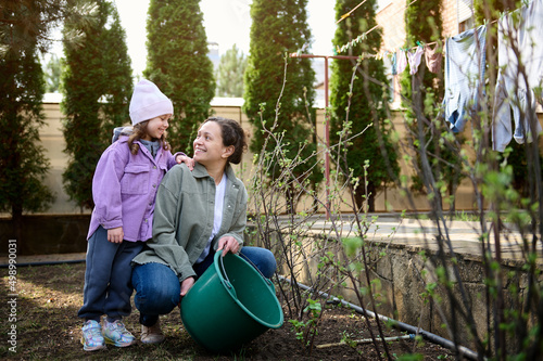 Happy mother and daughter watering flowering bushes and plants in the garden of the backyard of a country house, against the background of drying clothes on a rope on an early spring day