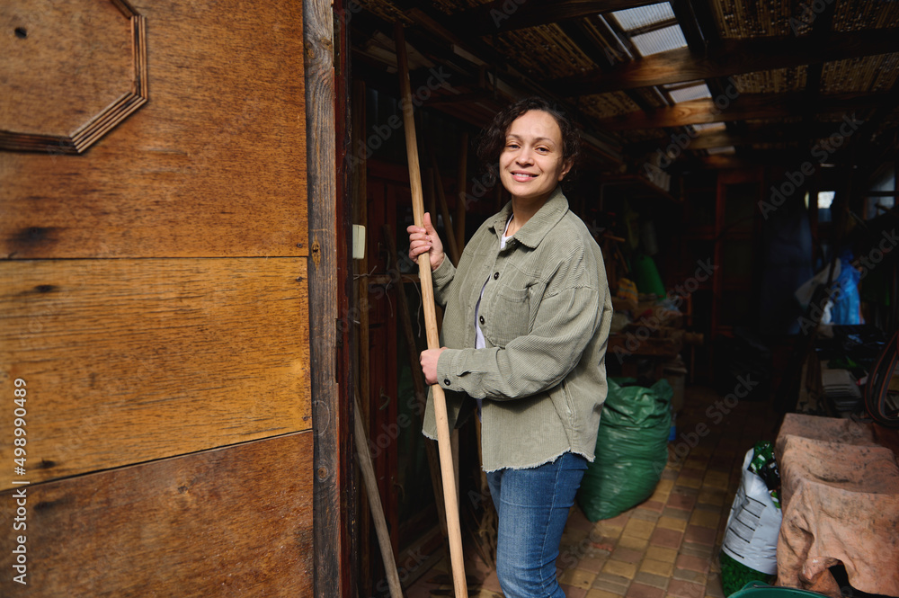 Happy female gardener farmer with gardening tools standing in her wooden rural workshop, enjoying gardening on a beautiful early spring day Happy female gardener farmer with gardening tools standing in her wooden rural workshop, enjoying gardening on a beautiful early spring day
