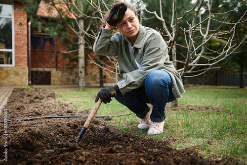 Busy female gardener in velvet shirt loosening the ground with a rake on a windy early spring day. Horticulture and gardening.