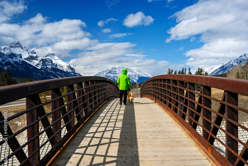 Unrecognizable people walking on a wood pedestrian bridge over cougar canyon in Canmore, with snowcapped mountain peaks against blue sky in the background