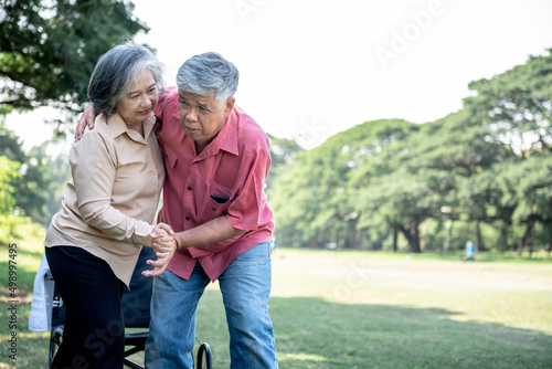 Asian elderly couple, attractive wife helping husband to get up from the wheelchair to do physical therapy with learning to walk, at green garden, to Asian couple and health care concept.