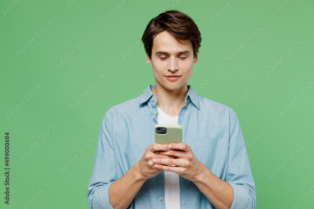 Dreamful pensive minded fancy young brunet man 20s years old wears blue shirt hold in hand use mobile cell phone typing browsing chatting send sms isolated on plain green background studio portrait. Dreamful pensive minded fancy young brunet man 20s years old wears blue shirt hold in hand use mobile cell phone typing browsing chatting send sms isolated on plain green background studio portrait.
