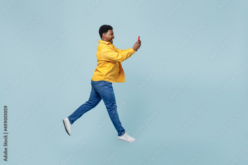 Full body side view young man of African American ethnicity 20s wear yellow shirt jump high run fast hold in hand use mobile cell phone isolated on plain pastel light blue background studio portrait