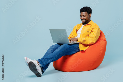 Full body smiling happy young man of African American ethnicity wear yellow shirt sit in bag chair hold use work on laptop pc computer isolated on plain pastel light blue background studio portrait