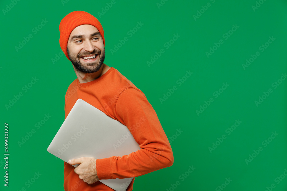 Side view young smiling happy man 20s in orange sweatshirt hat hold use work on laptop pc computer browsing internet look aside on workspace area copy space isolated on plain green background studio Side view young smiling happy man 20s in orange sweatshirt hat hold use work on laptop pc computer browsing internet look aside on workspace area copy space isolated on plain green background studio