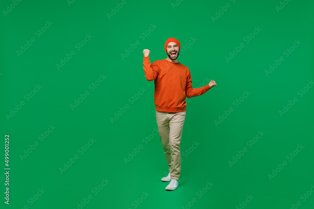 Full body young excited happy man 20s wear orange sweatshirt hat doing winner gesture celebrate clenching fists say yes isolated on plain green background studio portrait. People lifestyle concept Full body young excited happy man 20s wear orange sweatshirt hat doing winner gesture celebrate clenching fists say yes isolated on plain green background studio portrait. People lifestyle concept