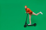 Full body side view young excited happy caucasian man 20s wear orange sweatshirt hat riding electric scooter raise up leg isolated on plain green background studio portrait. People lifestyle concept.