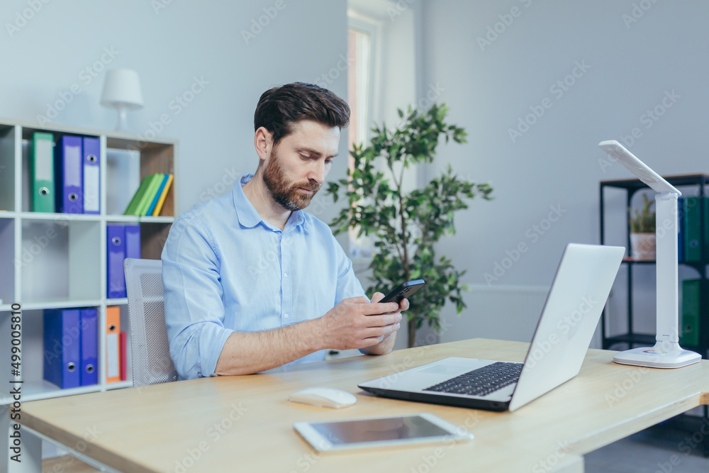 Fototapeta premium Focused young man with a beard, uses the phone, sitting at home at the table, thoughtfully reads from the screen