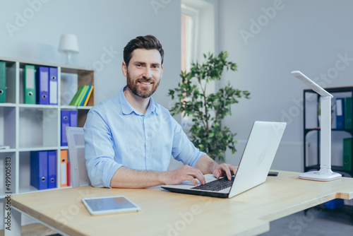 Happy and successful man working on laptop in home office, sitting at table in bright room, smiling looking at camera