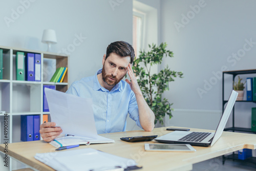 Pensive businessman freelancer at home, works at paperwork, examines thinking documents, husband works at home office at desk, uses laptop