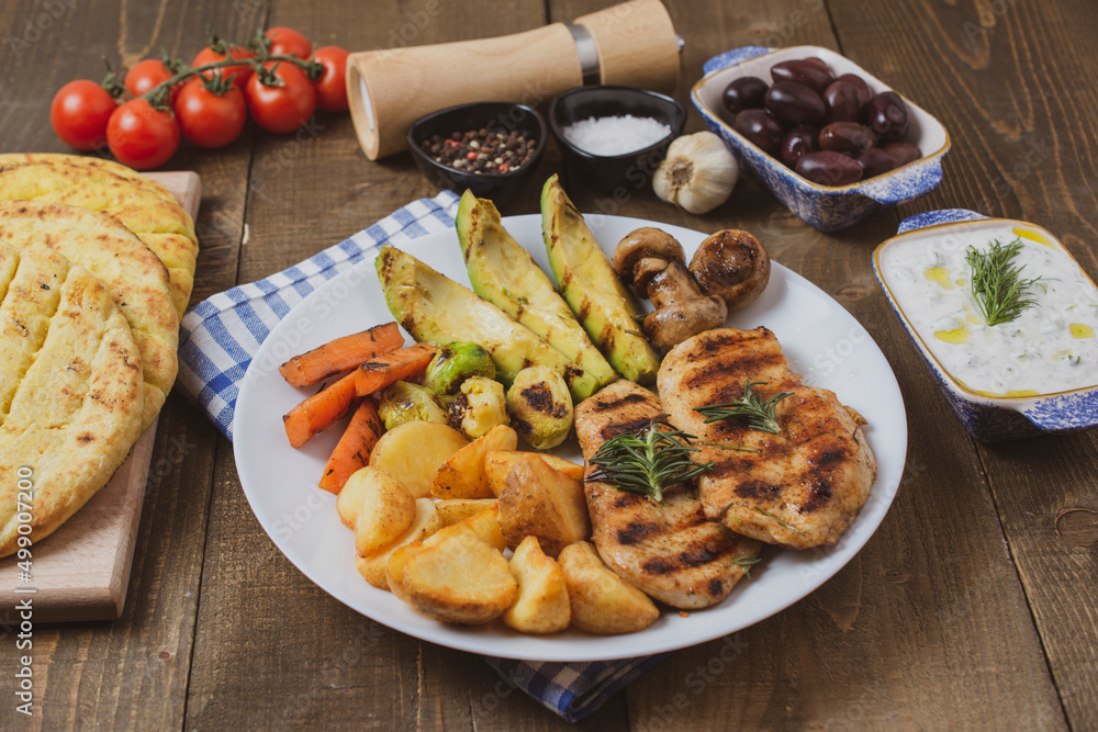 Tasty fresh meal with grilled chicken with potatoes, avocado, mushrooms, carrots in white bowl on wooden background. Olives, pita, tomatoes and tzatziki. Diet Keto Paleo Concept. Tasty fresh meal with grilled chicken with potatoes, avocado, mushrooms, carrots in white bowl on wooden background. Olives, pita, tomatoes and tzatziki. Diet Keto Paleo Concept.