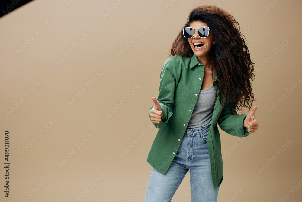 GOOD IDEA CONCEPT. Overjoyed smiling tanned curly Latin female in casual things sunglasses gesture thumbs up posing isolated over pastel beige background. Copy space Mockup Banner. Fashion offer