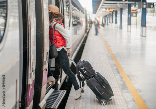 side view of a tourist with hand luggage, entering a train waiting at station. hat and red vest. concept weekend train trip. high speed train.