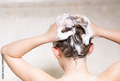 Little, beautiful girl, baby bathes sitting in a white bath with soap suds, shampoo and washes her head with hair. With your back to the camera