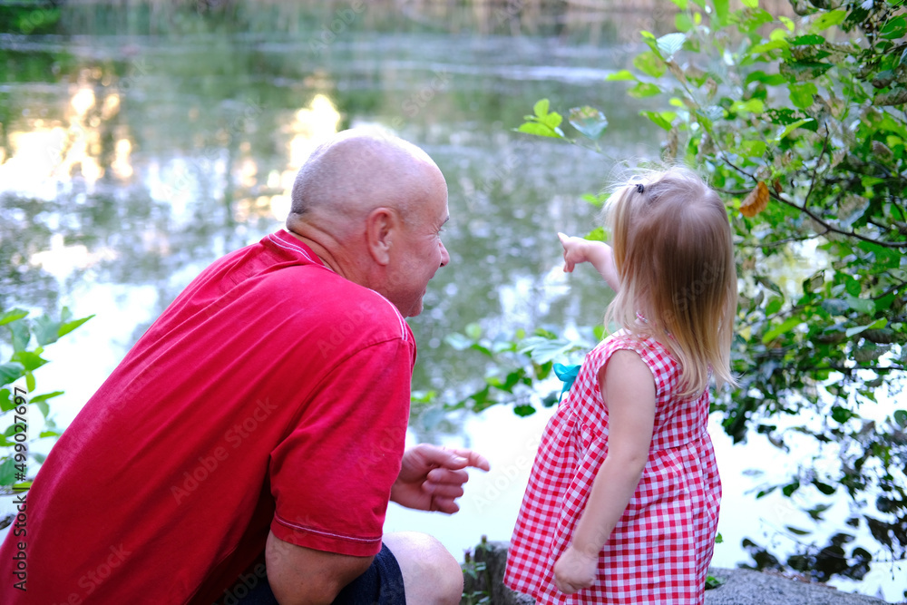 Naklejka premium small child, 2-year-old blonde girl in red dress, old man, grandfather and granddaughter by water, concept of childhood, family relationships of generations, walk in summer in nature