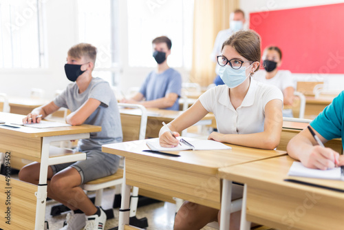 Teenagers in face masks sitting in class room. Male teacher explaining something to them.
