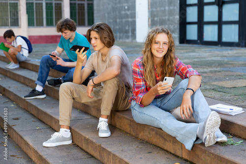 Teenage schoolers, boy and girl, using their gadgets outside school on sunny autumn day