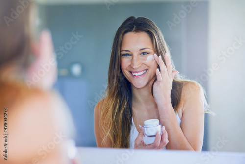 I moisturize daily for soft, smooth skin. Portrait of a beautiful young woman applying moisturizer to her face while looking in the mirror. photo