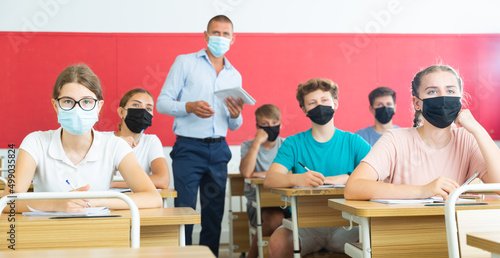 Focused teenage students in protective face masks studying in classroom with teacher, writing lectures in workbooks. Necessary precautions in coronavirus pandemic