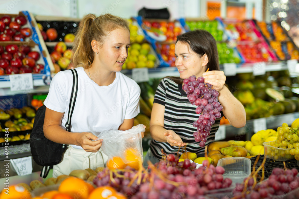 Young woman with a fifteen-year-old girl, who came to the store for shopping, choose a bunch of grapes, holding a package of ..oranges in her hands