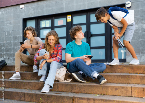 Teenager girl and boys sitting front college with smartphones in hands.
