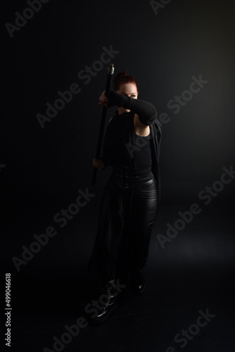 Full length portrait of pretty redhead female model wearing black futuristic scifi leather cloak costume. Standing pose on dark studio background with shadow rim  moody lighting.