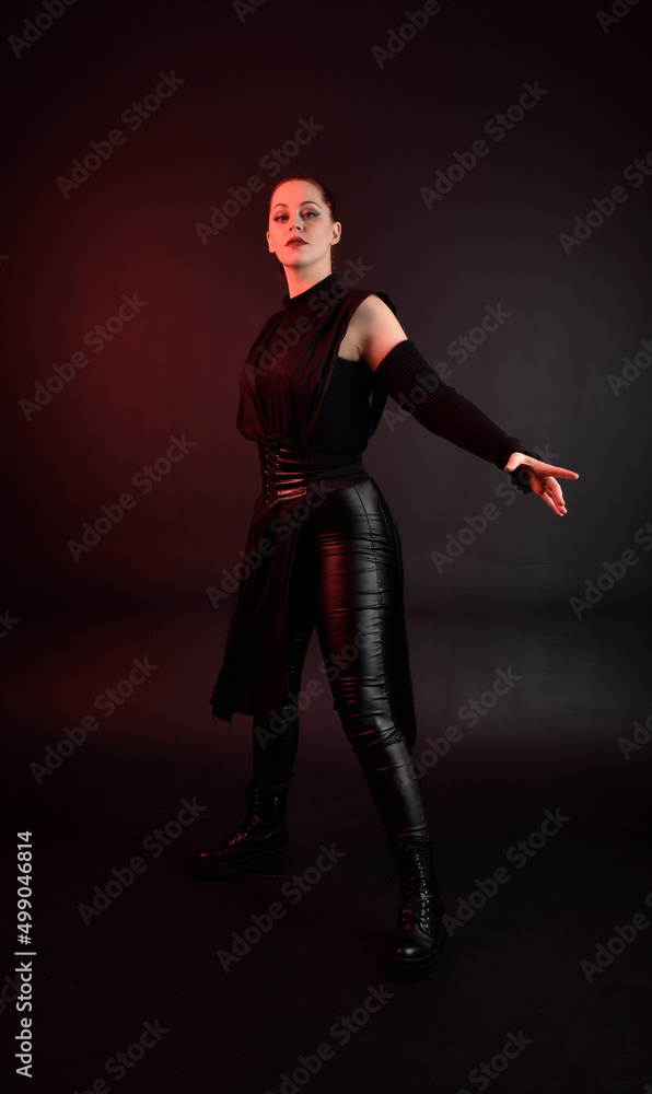 Full length portrait of pretty redhead female model wearing black futuristic scifi leather cloak costume. Standing pose on dark studio background with shadow rim  moody lighting.