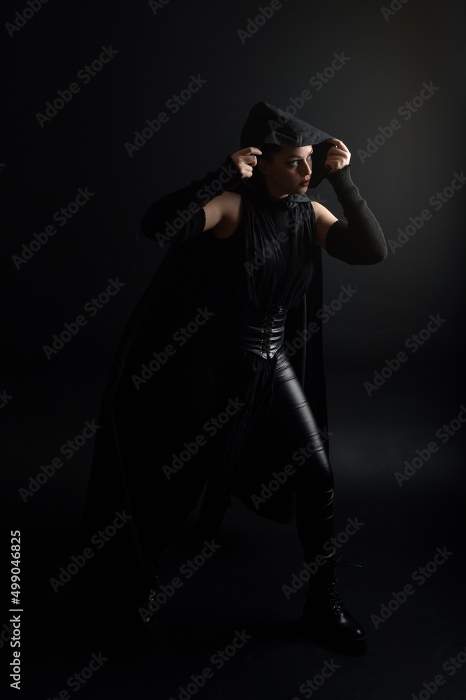 Full length portrait of pretty redhead female model wearing black futuristic scifi leather cloak costume. Standing pose on dark studio background with shadow rim moody lighting. Full length portrait of pretty redhead female model wearing black futuristic scifi leather cloak costume. Standing pose on dark studio background with shadow rim moody lighting.