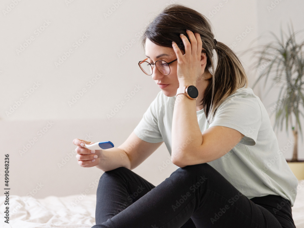 the woman found out about an unwanted pregnancy. A woman with a pregnancy test in her hands is sitting on the bed in her apartment - obrazy, fototapety, plakaty 