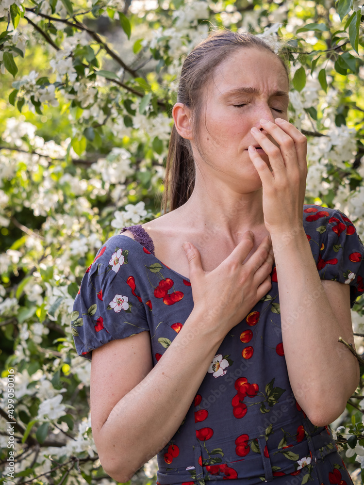 Young woman sneezes in the park against the background of a flowering tree. Allergy to pollen concept. Young woman sneezes in the park against the background of a flowering tree. Allergy to pollen concept.