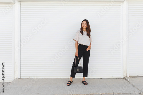 Young beautiful woman, wearing beige t-shirt, black pants, bag with chain and flat sandals walking outdoor near white roller door. Stylish trendy basic minimalistic casual outfit. Street fashion. photo
