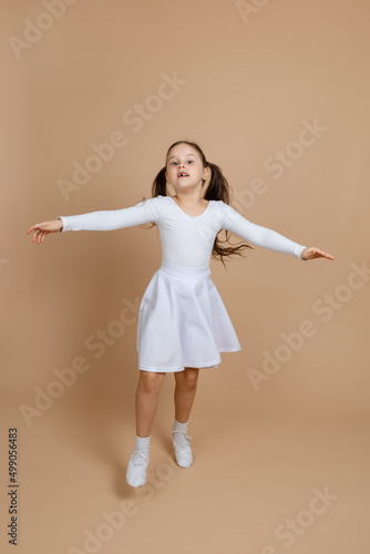 Young cute girl with long dark hair in white dress, socks and gymnastics shoes whirling, dancing, spreading hands, raising leg, having fun on brown background. Ballet, gymnastics, sport, cheerleading.