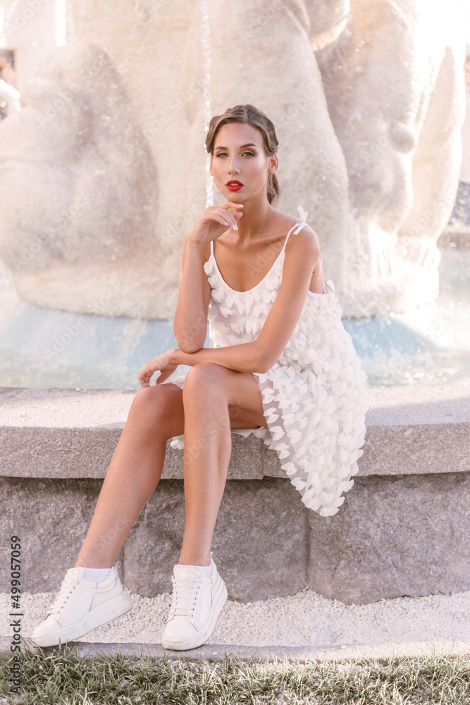 A stylish bridesmaid in a white light dress sits by a water fountain. Lovely make-up and fashion hairstyle
