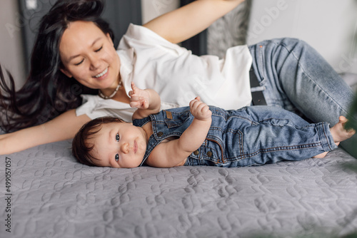 Portrait of smiling mother with little daughter in bedroom closeup, free copy space, blurred background. Cozy lay on bed with baby, capture happy emotions. Concept of maternal affection and childcare. © Юля Бурмистрова