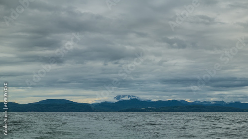 Kamchatka coast against a cloudy sky. A snow-covered volcano hides in low clouds. Ripples on the surface of the Pacific Ocean. Avacha Bay