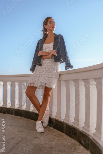 An attractive woman celebrates life in a summer white dress and denim jacket on the balcony of a summer resort. Joy and serenity