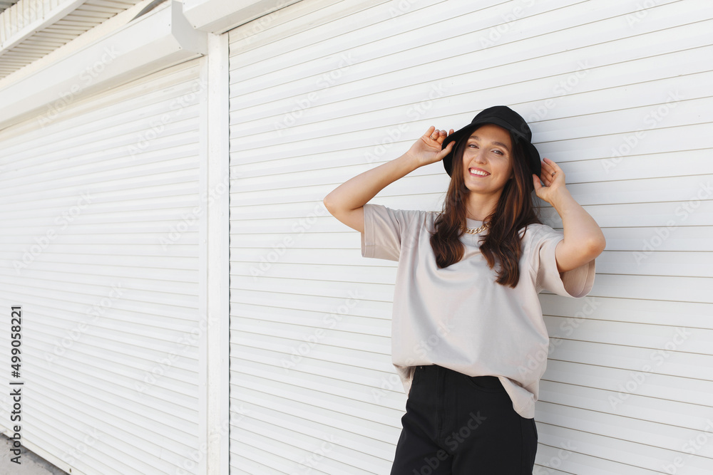 Young beautiful woman, wearing beige t-shirt, black pants, bucket hat and gold chain necklace, standing outdoor near white roller door. Stylish trendy basic minimalistic casual outfit. Street fashion. Young beautiful woman, wearing beige t-shirt, black pants, bucket hat and gold chain necklace, standing outdoor near white roller door. Stylish trendy basic minimalistic casual outfit. Street fashion.