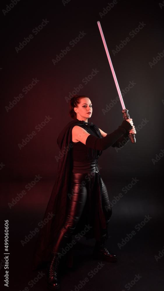 Full length portrait of pretty redhead female model wearing black futuristic scifi leather cloak costume. Standing pose holding lightsaber on dark studio background with shadow moody lighting. Full length portrait of pretty redhead female model wearing black futuristic scifi leather cloak costume. Standing pose holding lightsaber on dark studio background with shadow moody lighting.