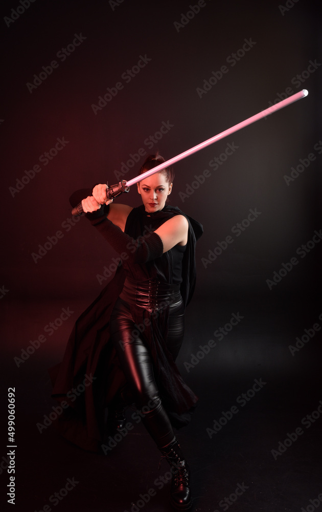 Full length portrait of pretty redhead female model wearing black futuristic scifi leather cloak costume. Standing pose holding lightsaber on dark studio background with shadow moody lighting. Full length portrait of pretty redhead female model wearing black futuristic scifi leather cloak costume. Standing pose holding lightsaber on dark studio background with shadow moody lighting.