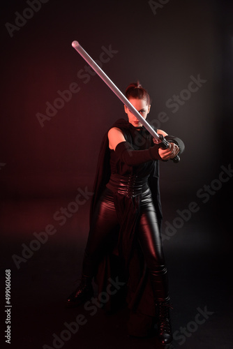 Full length portrait of pretty redhead female model wearing black futuristic scifi leather cloak costume. Standing pose holding lightsaber on dark studio background with shadow moody lighting.