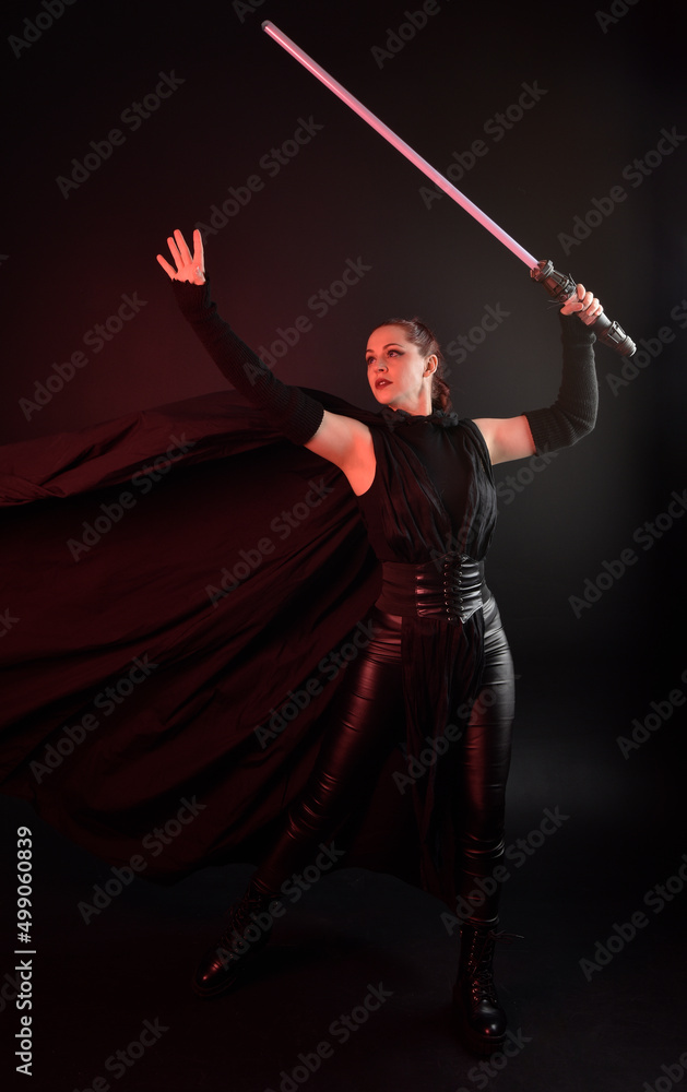 Full length portrait of pretty redhead female model wearing black futuristic scifi leather cloak costume. Standing pose  holding lightsaber on dark studio background with shadow moody lighting.
