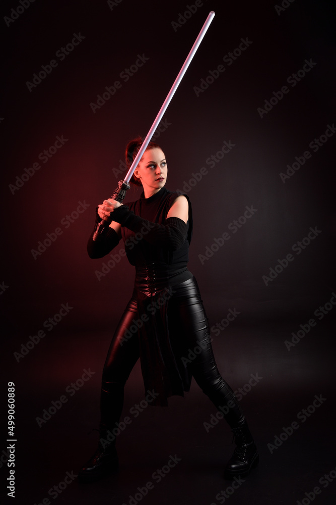 Full length portrait of pretty redhead female model wearing black futuristic scifi leather cloak costume. Standing pose holding lightsaber on dark studio background with shadow moody lighting. Full length portrait of pretty redhead female model wearing black futuristic scifi leather cloak costume. Standing pose holding lightsaber on dark studio background with shadow moody lighting.
